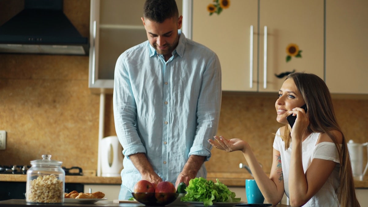 Attractive couple in the kitchen early morning. Beautiful girl chatting phone while her boyfriend cooking breakfast at home